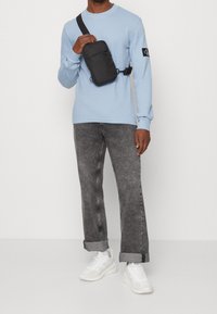 Muže v bleděmodré košili s dlouhým rukávem, s černou crossbody taškou, šedých jeansových kalhotách s ohrnutými nohavicemi a bílých teniskách, stojícího na bílém pozadí.