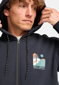 Sudadera con cremallera de color marino con un acabado texturizado, que presenta una impresión gráfica de un paisaje, cordones con acentos dorados y capucha forrada.