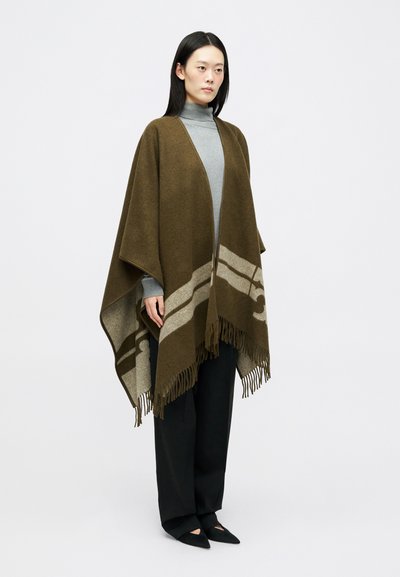 Poncho de lã castanho com bordas em franjas e um padrão abstrato em castanho mais claro. Usado sobre um suéter de gola alta cinza e combinado com calças pretas.