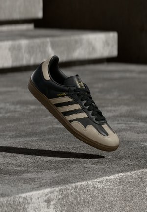 Baskets Adidas Samba noires et beiges en l'air au-dessus d'escaliers en béton, montrant le côté avec trois bandes et une semelle en gomme en extérieur.