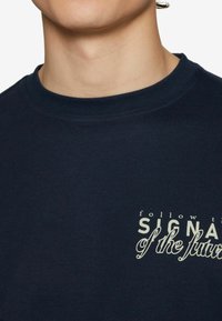 Námorně modré bavlněné tričko s kulatým výstřihem, na kterém je krémovým písmem napsáno "následuj SIGNÁL budoucnosti" přes hrudník.