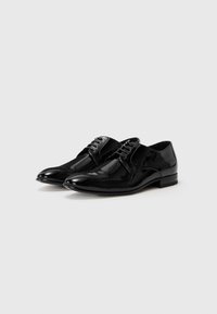 Un par de zapatos de vestir de cuero negro brillante con cordones sobre un fondo gris claro liso, en ángulo hacia la izquierda.