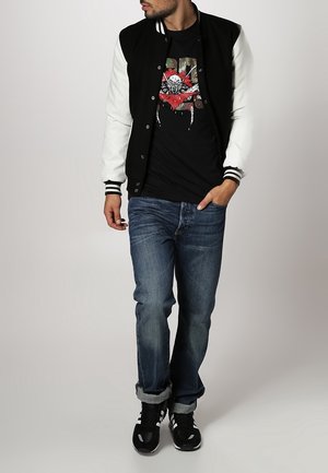 Giacca varsity bianca e nera con bottoni a scatto, indossata sopra una maglietta grafica nera, abbinata a jeans blu arrotolati e scarpe da ginnastica nere.