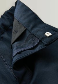 Mørkeblå stof med en struktureret finish, udstyret med en lynlåslukning og en metalknapdetalje ved linningen. Indvendig foring synlig.