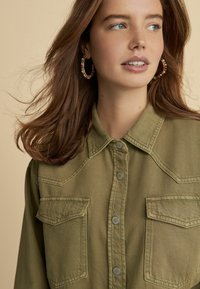 Olive zelená denimová košeľa s dvoma gombíkovými náprsnými vreckami, patentnými gombíkmi a klasickým golierom. Modelka má jemné, splývavé vlasy.