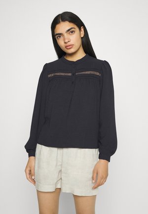 Blusa de manga longa azul-marinho com um colarinho alto, apresentando detalhes em renda no peito e fecho de botão, combinada com calções de linho leves.
