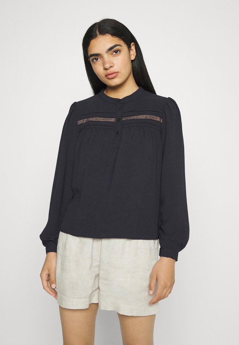 Blusa de manga longa azul-marinho com um colarinho alto, apresentando detalhes em renda no peito e fecho de botão, combinada com calções de linho leves.