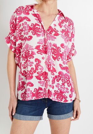 Donna che indossa una camicetta bianca con motivi floreali rosa acceso e pantaloncini di jeans blu scuro risvoltati.