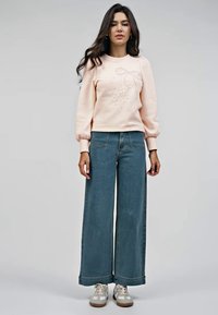 Lys rosa genser med pufferm og dekorativ sløyfemønster, sammen med brede blå jeans og nøytrale joggesko. Enkelt design.