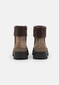 Brązowe zamszowe botki za kostkę z dzianinowym brązowym mankietem, gumową podeszwą i tłoczonym wzorem logo. Widok z tyłu podkreśla fakturowaną cholewkę i solidną konstrukcję.