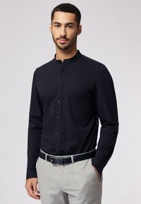 Donkerblauwe knoopshirt met lange mouwen en staande kraag, gecombineerd met lichtgrijze broek en een donkere riem, met een zachte stoftextuur.
