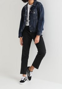 Giacca di jeans blu scuro con bottoni argentati, T-shirt bianca sotto, pantaloni neri a pochi centimetri dalla caviglia e sneakers nere con lacci bianchi.