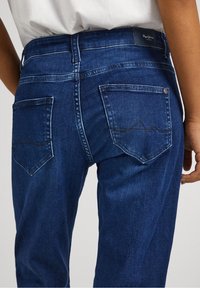 Modré džíny ze silné bavlny mají rovný střih, kontrastní prošívání, dvě zadní kapsy s vlnkovaným vzorem a značenou páskou na elastickém opasku.