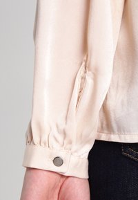 Blusa satinata color pesca chiaro con polsini a manica arricciati e dettaglio di bottoni argentati; texture liscia e leggera lucentezza, abbinata a jeans scuri.