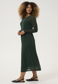 Green, long-sleeve dress with a fitted bodice and flowy skirt, made of a shimmery, textured fabric. Paired with black, flat sandals featuring accents.