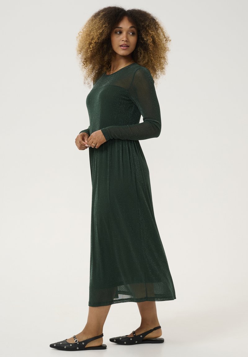Green, long-sleeve dress with a fitted bodice and flowy skirt, made of a shimmery, textured fabric. Paired with black, flat sandals featuring accents.