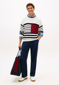 Λευκό και ναυτικό ριγέ πουλόβερ με κόκκινη λεπτομέρεια, φορεμένο πάνω από ένα ανοιχτό μπλε πουκάμισο με γιακά. Σκούρο μπλε παντελόνι και ναυτικό τσάντα ώμου με κόκκινο πάνελ.