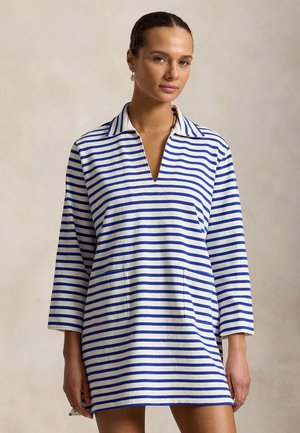 Kvinde iført en langærmet hvid kjole med blå vandrette striber og V-hals, stående foran en ensfarvet beige baggrund.