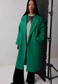 Roheline kahekordne ülikond, mis on valmistatud paksust materjalist laia kraega, koos halli sviitriga all ja mustade laiade pükstega.