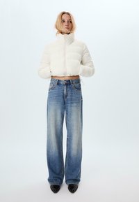 Vit kort pufferjacka med hög krage och frontdragkedja, i kombination med ljusblå vida jeans och svarta spetsiga skor.