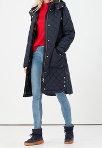 Donna che indossa un lungo cappotto trapuntato blu navy su un maglione rosso, jeans skinny azzurri chiari e stivali alla caviglia blu navy con lacci e suole beige.