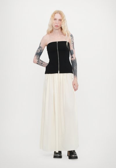 Svart strapless topp med silverdragkedja, tillsammans med en krämfärgad, plisserad maxikjol. Modellen bär svarta platåskor och visar upp tatuerade armar.