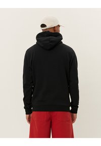 Felpa nera con cappuccio e polsini e orlo a coste, mostrata al contrario. Abbinata a pantaloni in velluto a coste rossi. Design casual, materiale in cotone.