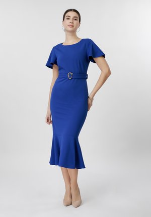 Woman in a fitted royal blue dress with short sleeves, belted waist, flared hem, beige heels, and hoop earrings standing against white background.