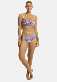 Das Model posiert in einem floralen Bikini vor einem hellblauen Hintergrund, mit einem entspannten Ausdruck und barfuß.