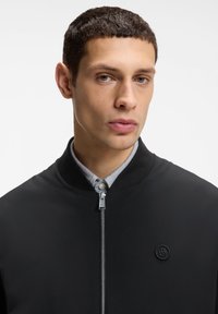 Νέος άνδρας με κοντά σκούρα μαλλιά, φορώντας μαύρο μπουφάν με φερμουάρ πάνω από ανοιχτό γκρι πουκάμισο με γιακά, σε λευκό φόντο.