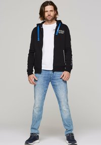 Sudadera negra con cremallera y cordones azules, gráfico blanco en el pecho, combinada con jeans de color azul claro y zapatillas oscuras.