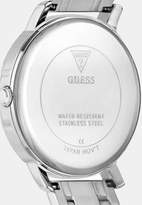 Edelstahl-Uhr mit silberner Farbe, mit einem runden Gehäuseboden, der mit "GUESS", "WASSERDICHT" und "JAPAN MOV'T" graviert ist.