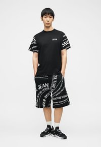 Νέος άνδρας στέκεται φορώντας ένα μαύρο και άσπρο σύνολο Versace Jeans Couture με τυπωμένο κείμενο σε σορτς, μανίκια πουκάμισου και αθλητικά παπούτσια.