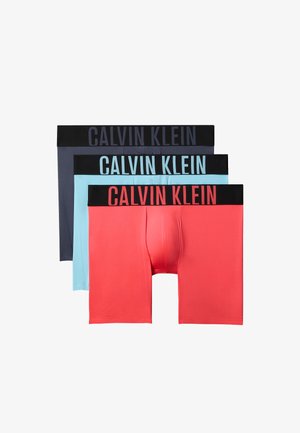 Jeune homme noir debout pieds nus, portant un caleçon ajusté Calvin Klein gris foncé, une main reposant sur sa hanche.