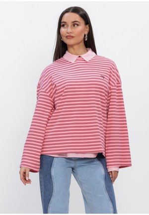 Vrouw draagt een losse roze gestreepte lange mouwen blouse met kraag en blauw twee-kleurige jeans, staande tegen een witte achtergrond.