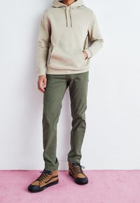 Beige mikina s kapucí a přední kapsou, olivově zelené kalhoty a hnědé šněrovací boty s černou podrážkou, umístěné na růžovém pozadí.