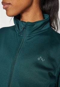 Giacca zip-up di un verde scuro realizzata in un tessuto liscio e stretch, con colletto alto e un sottile dettaglio del logo sul petto.