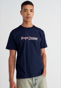 Tums zila kokvilnas blūze ar īsām piedurknēm, ar tekstu "Pepe Jeans Portobello Rd W1" gaiši rozā krāsā, apaļu kakla izgriezumu un brīvu piegriezumu.