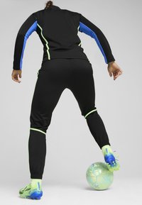 Zwart sporttrainingspak met blauwe en neon groene accenten, aansluitend ontwerp en bijpassende groene sportschoenen. Een speler raakt een bal met één voet aan.