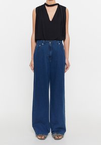 Top negro sin mangas con escote en V profundo y tela drapeada, combinado con jeans de tiro alto y pierna ancha en azul, que cuentan con bolsillos frontales.