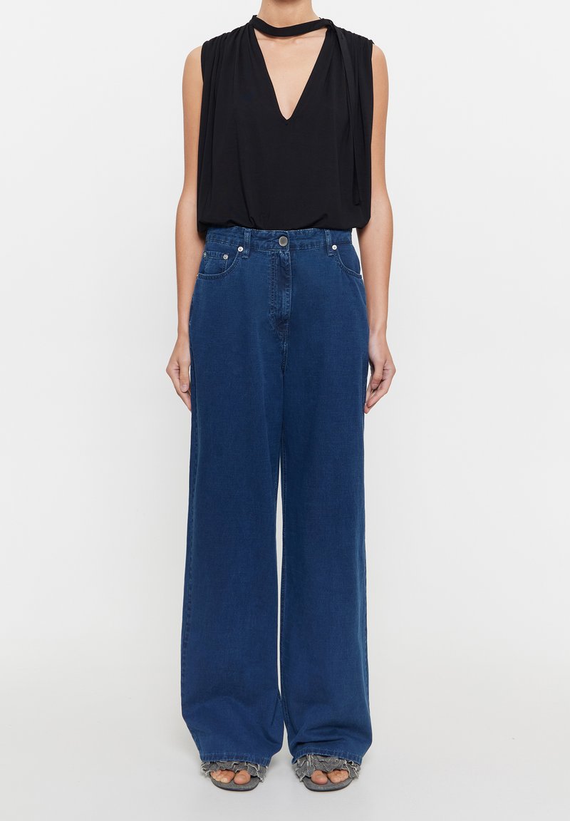 Top negro sin mangas con escote en V profundo y tela drapeada, combinado con jeans de tiro alto y pierna ancha en azul, que cuentan con bolsillos frontales.