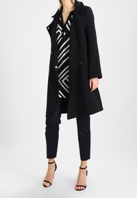 Schwarzer Wollmantel mit Reverskragen, kombiniert mit einem schwarz-weiß gestreiften Hemd und marineblauen, strukturierten Hosen, ergänzt durch schwarze, geschnallte High Heels.