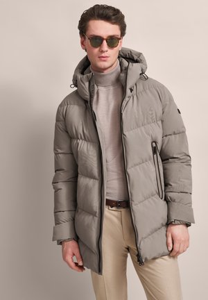 Puffjacke in Hellgrau mit Kapuze, gestepptem Muster, Reißverschlusstaschen und einem schlanken Design. Über einem beigen Rollkragenpullover getragen.