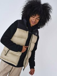 Gilet imbottito beige con yoke nero, dotato di una zip completa e due tasche frontali. Indossato sopra una felpa nera con cappuccio.