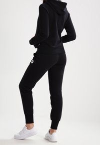 Sweat à capuche noir et joggings assortis en tissu doux. La tenue est dotée de poignets côtelés et d'un écusson logo sur le pantalon. Des baskets blanches complètent le look.