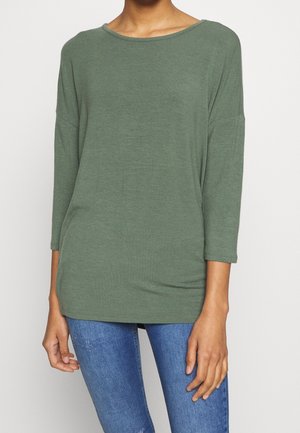 Osoba ubrana w luźną, oliwkowozieloną bluzkę z długim rękawem i okrągłym dekoltem oraz niebieskie dżinsy, stojąca na jednolitym tle.