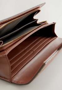 Portefeuille en cuir marron avec des fermetures éclair dorées, plusieurs compartiments et emplacements pour cartes. L'intérieur présente une doublure noire et des détails cousus.