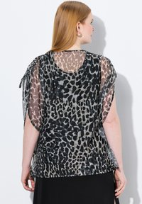 Blouse transparente en noir, gris et imprimé léopard beige. Présente une coupe ample, des manches courtes et un col rond. Des fentes sur les côtés ajoutent du détail.