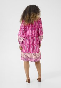 Roza obleka z vzorcem paisley, sproščen kroj, dolgi rokavi, zategnjen pas in tekoč rob. Obuta v sandale z odprto konico.