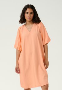 Perzikkleurige jurk met een V-hals en pofmouwen. Gemaakt van een lichtgewicht stof, met een losse, comfortabele pasvorm.